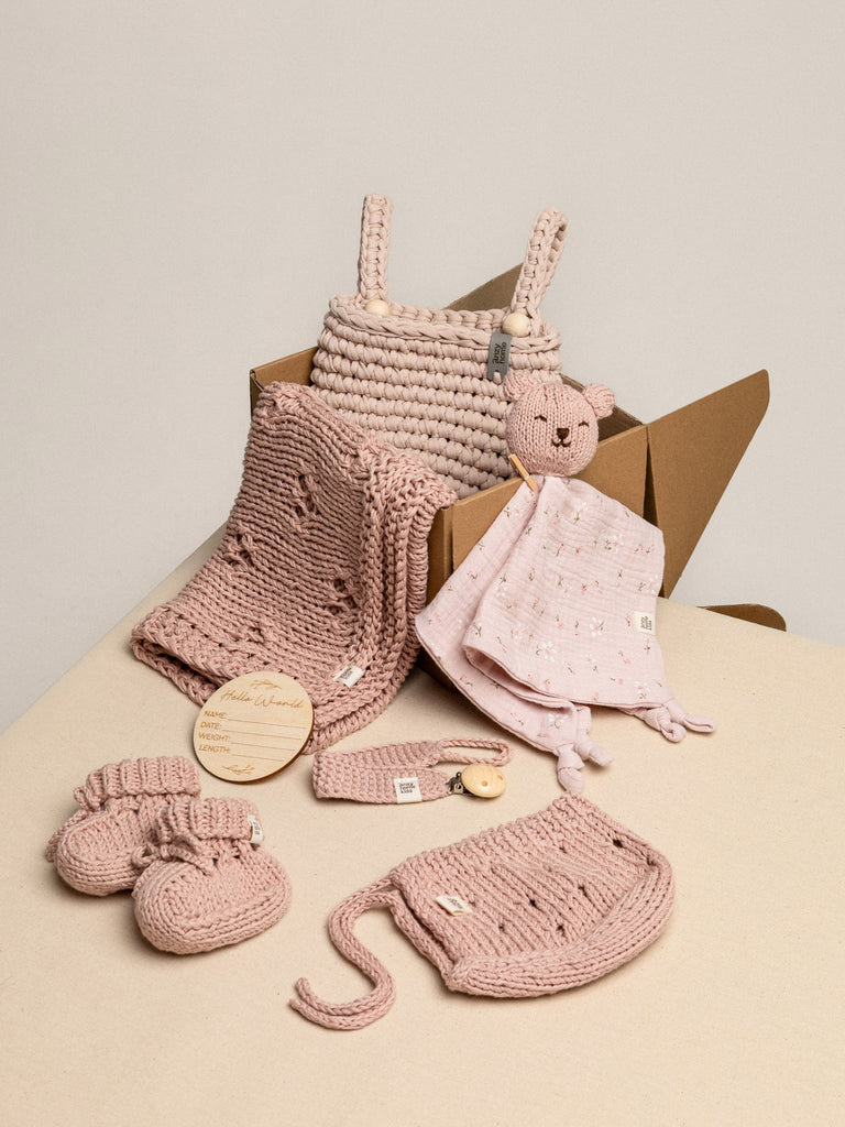 Collection of pink baby items including a knitted blanket, toy, and booties on a beige surface.
